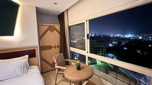 A hotel room with a bed, a chair, a small table with a flower, and a large window offering a city view at night. - Grand Museum Guest House pyramids view (Giza)