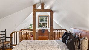 A bedroom with a sloped ceiling, wooden beams, a window, a bed, a chair, and a small balcony. - Ocean Views from 2 Decks! Beachfront ⎮ Amphitrite (Ucluelet)