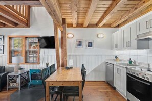 A kitchen with wooden beams, white cabinets, a dining table, and a TV mounted on the wall. - Ocean Views from 2 Decks! Beachfront ⎮ Amphitrite (Ucluelet)