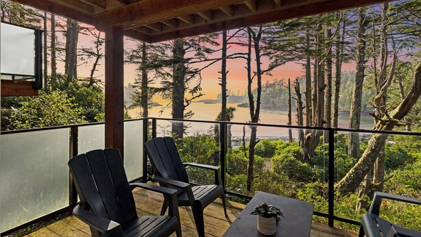 A wooden deck with two Adirondack chairs and a small table, overlooking a forested area and a river. - Ocean Views from 2 Decks! Beachfront ⎮ Amphitrite (Ucluelet)