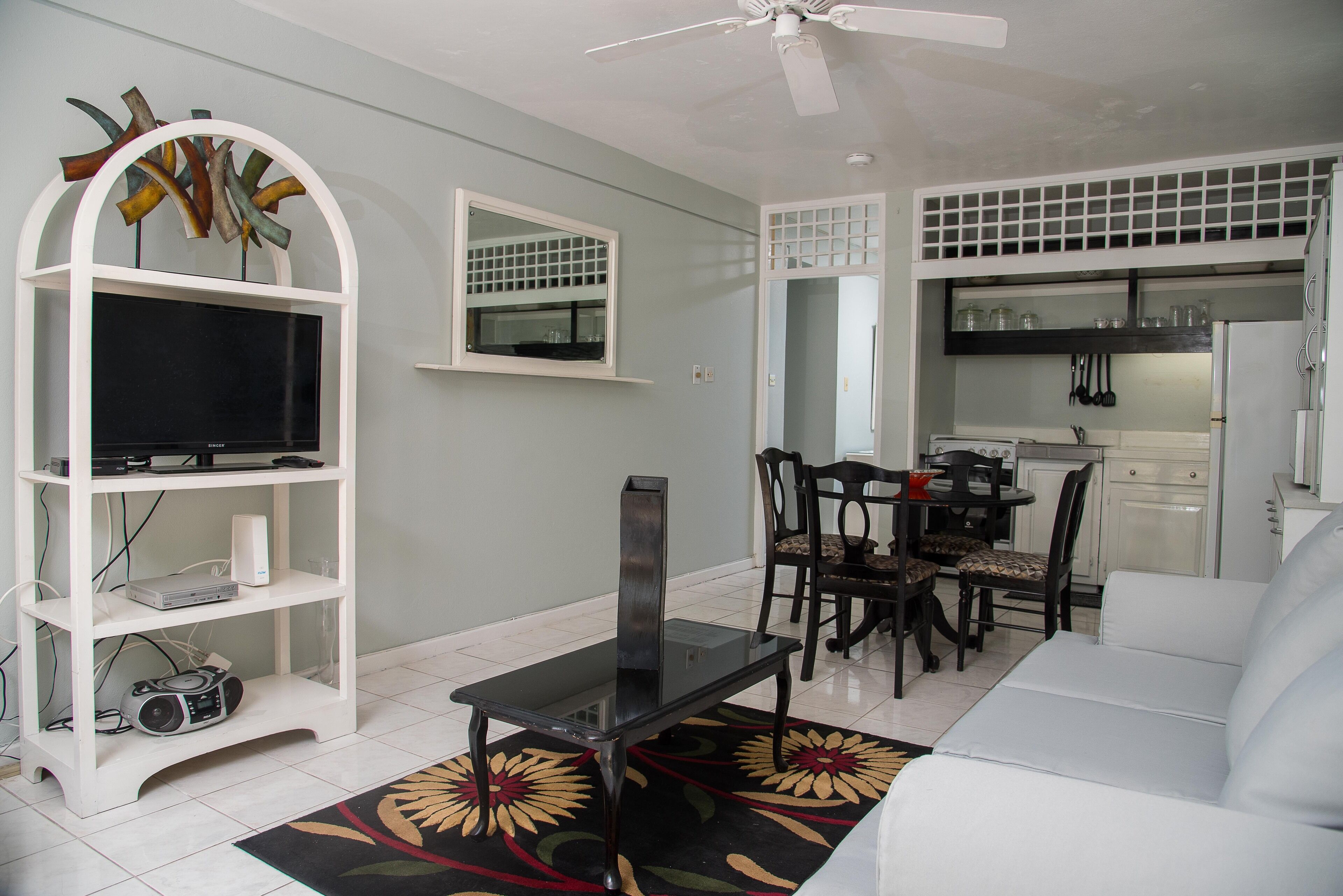 Uma sala de estar moderna com televisão de tela plana, uma mesa de jantar com cadeiras, um sofá e uma área de cozinha com armários e geladeira.