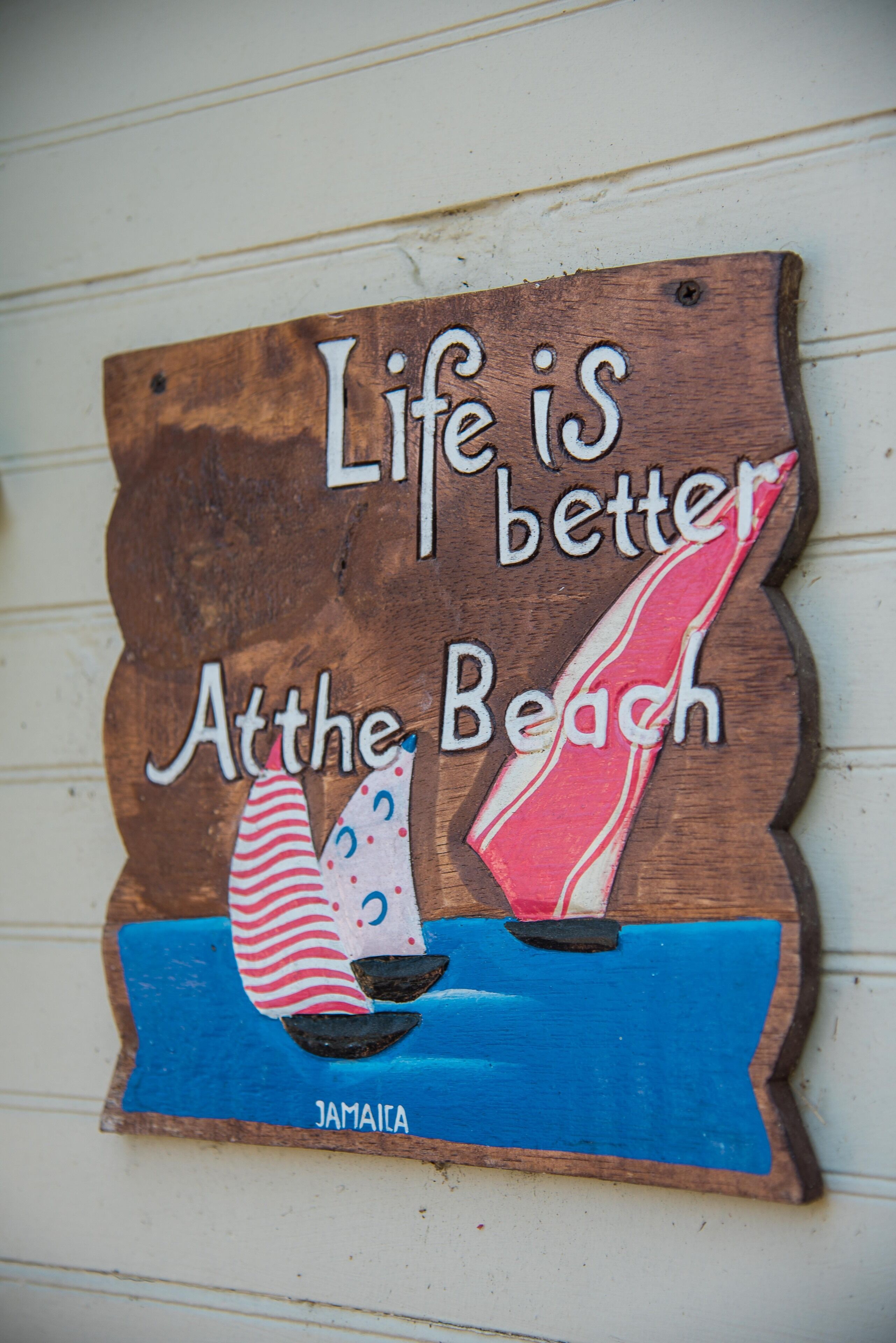 Un letrero de madera con el texto 'Life is better at the beach' e ilustración de un velero.