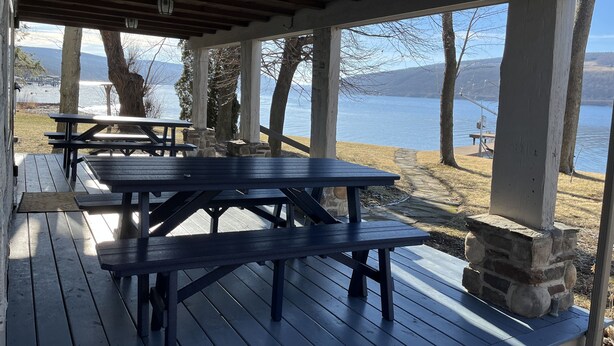 An outdoor covered picnic area with wooden benches and tables, stone pillars, and a view of a lake and mountains. - Moonlight Manor on Keuka Lake in Hammondsport (Hammondsport)