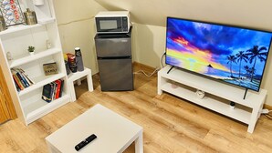 A compact living space with a flat-screen TV, a coffee machine, a mini-fridge, a white coffee table, and a bookshelf with books and decorative items. - Bright, Stylish & Cozy Private Attic Unit near NYC (HALEDON)