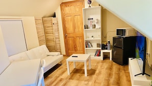 A cozy living room with a white sofa, a small white table, a wooden wardrobe, a bookshelf, a black refrigerator, and a microwave on a shelf. - Bright, Stylish & Cozy Private Attic Unit near NYC (HALEDON)