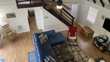 Un vaste salon comprenant un canapé d’angle bleu, un fauteuil à bascule en bois, un fauteuil rouge et une petite table sur laquelle se trouve un panier. On peut apercevoir un ventilateur de plafond, et un escalier mène à un niveau supérieur.