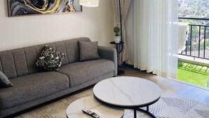 Una sala de estar moderna con un sofá gris, dos mesas redondas, una araña, una lámpara de pie, un cuadro y vistas de un paisaje urbano a través de grandes ventanales.