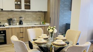 Comedor con una mesa redonda dispuesta para seis personas, rodeada de sillas tapizadas con detalles dorados. Un candelabro moderno cuelga encima, y un jarrón con flores blancas está sobre la mesa.