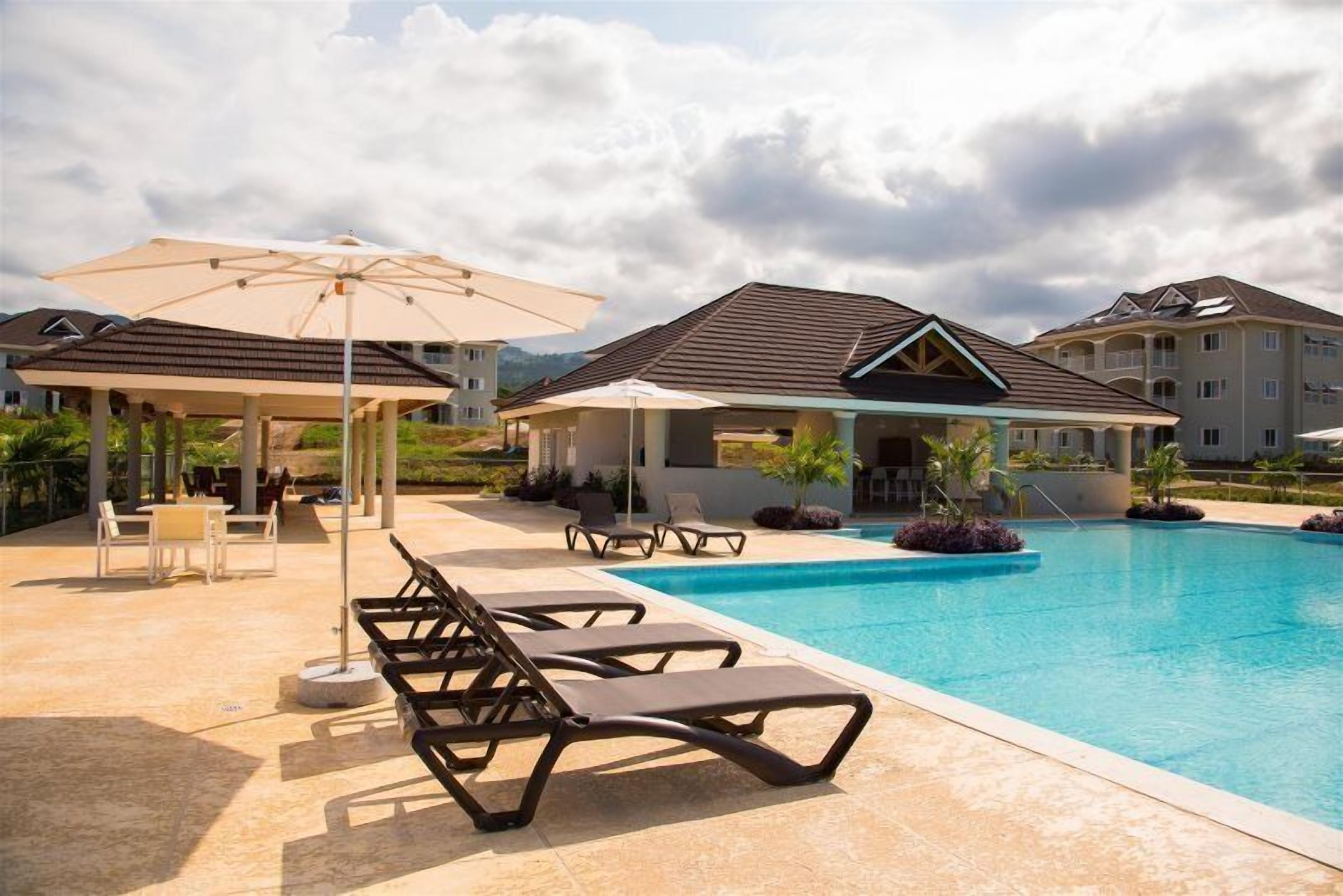 Área de piscina com espreguiçadeiras, guarda-sóis e um espaço coberto para sentar.