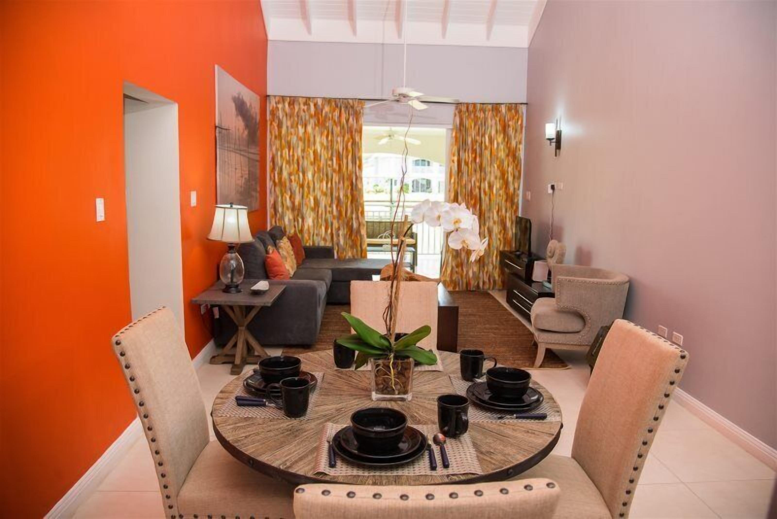 A dining area with a round table set for four, surrounded by upholstered chairs. A small table with a lamp and a vase with an orchid is visible in the background.