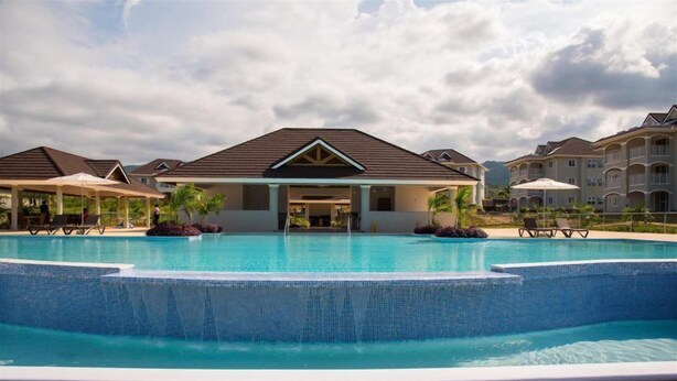A swimming pool with a wooden deck, surrounded by buildings with a central entrance and covered seating areas. - Imperial Topaz At Richmond (St. Ann's Bay)