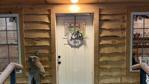 En entré till en stuga med en vit dörr, en skylt med texten ”Här är det gott att vara hemma” och en träskulptur av en uggla.