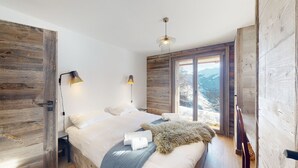 Un dormitorio con una cama grande, paredes de madera, dos lámparas de noche y vistas a montañas nevadas a través de un ventanal corredera.