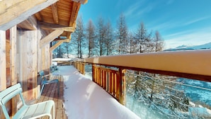 Una terraza de madera con suelo nevado y vistas a árboles y montañas cubiertas de nieve.