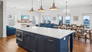 A spacious kitchen with a large island, wooden cabinets, and pendant lighting. There are multiple windows offering views of the ocean.