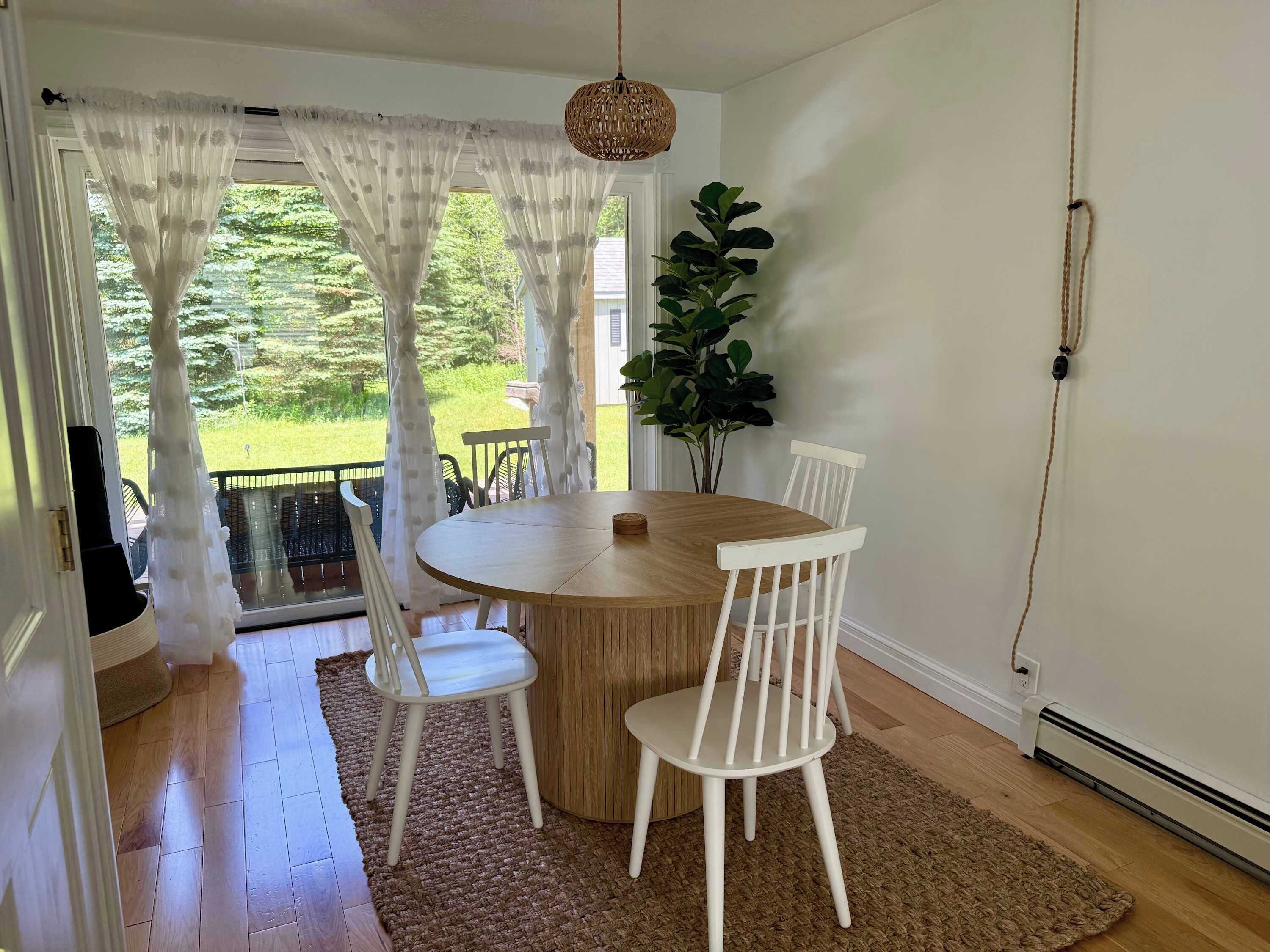 A dining area with a round table, white chairs, a woven pendant light, and a view of a green landscape through sheer curtains.
