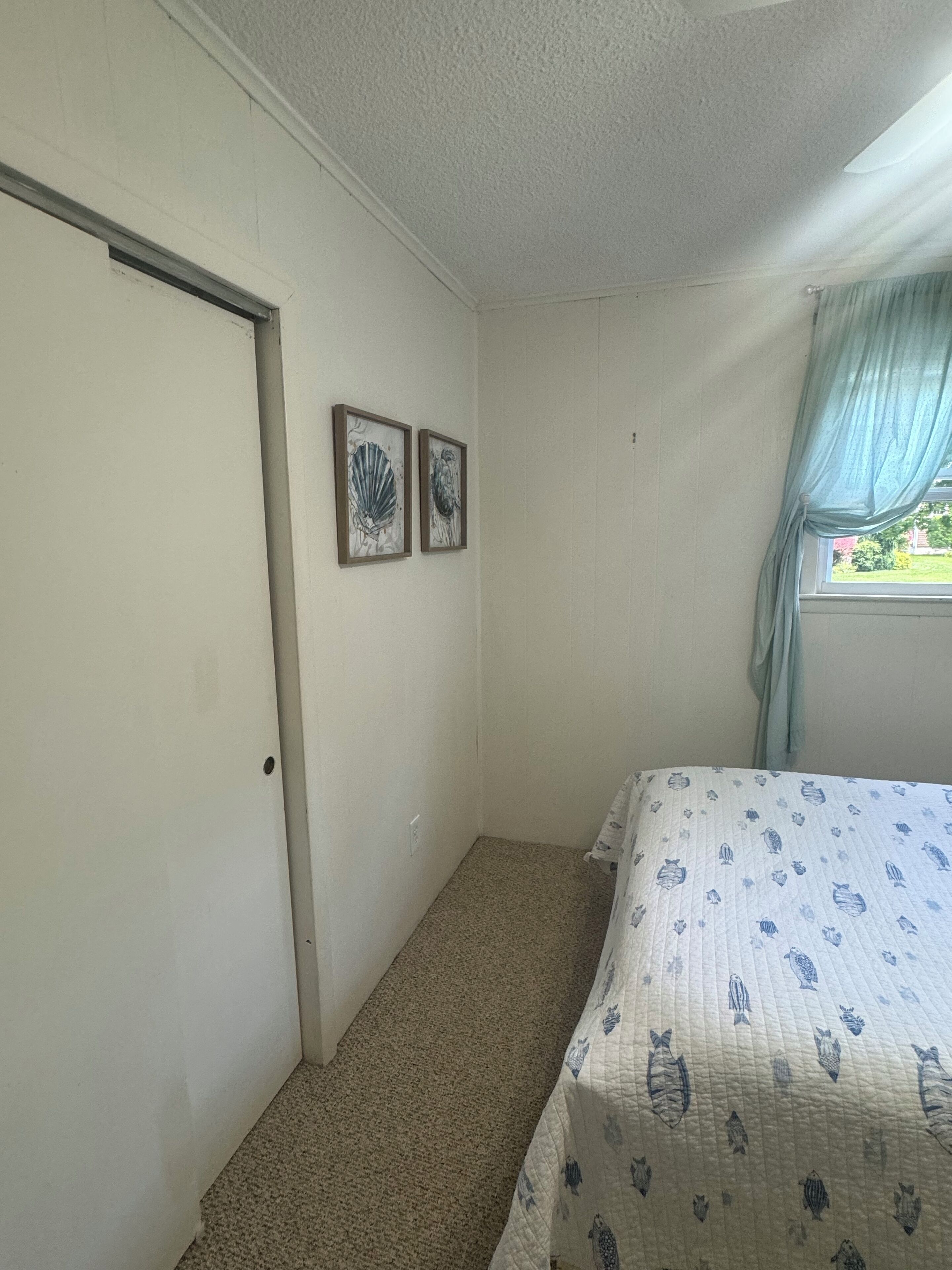 A small, sparsely furnished room with a single bed, a door, and two framed artworks on the wall.