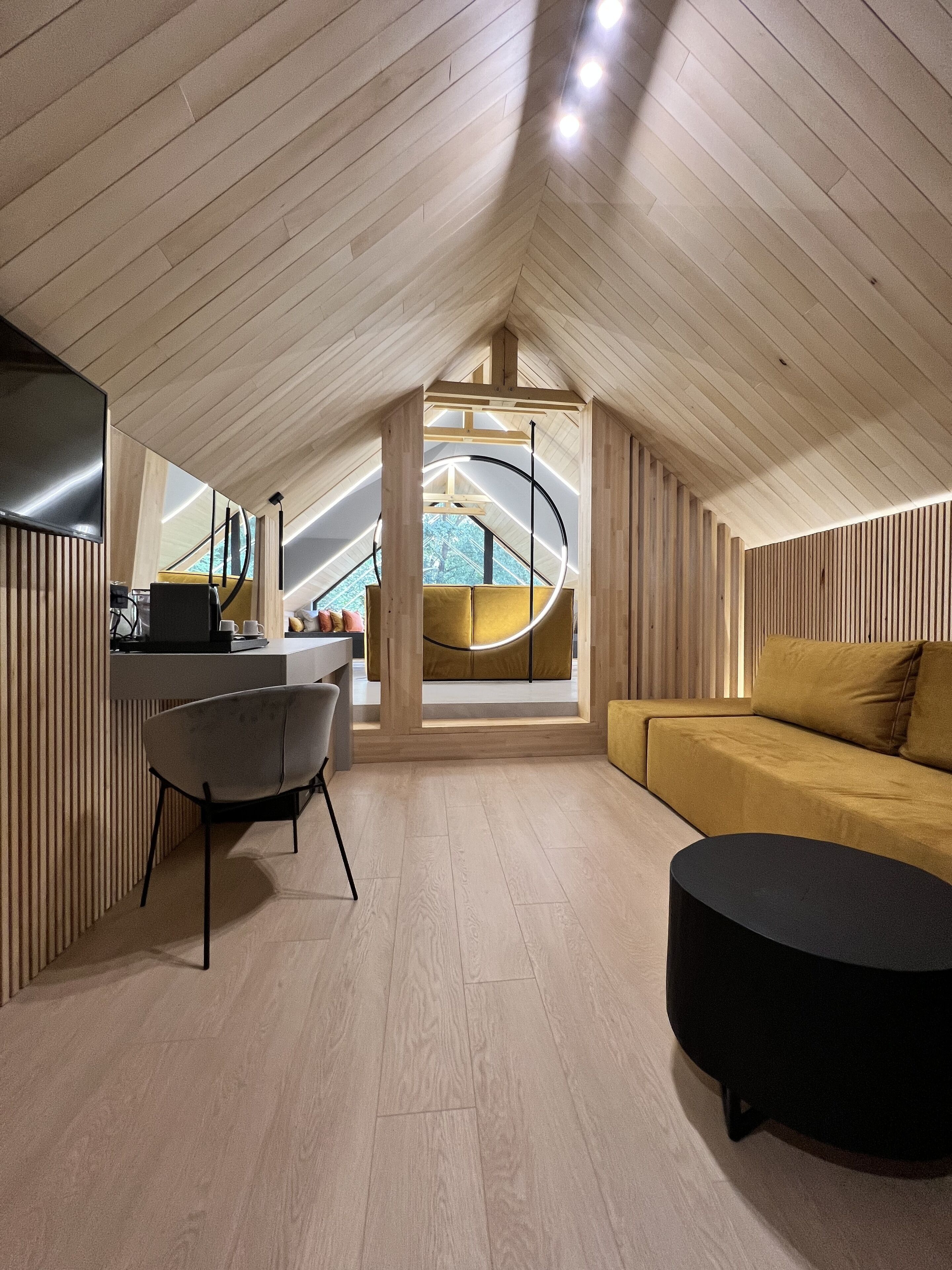 A modern loft bedroom with a sloped ceiling, a central circular light fixture, a sofa, a dining table, and a TV.