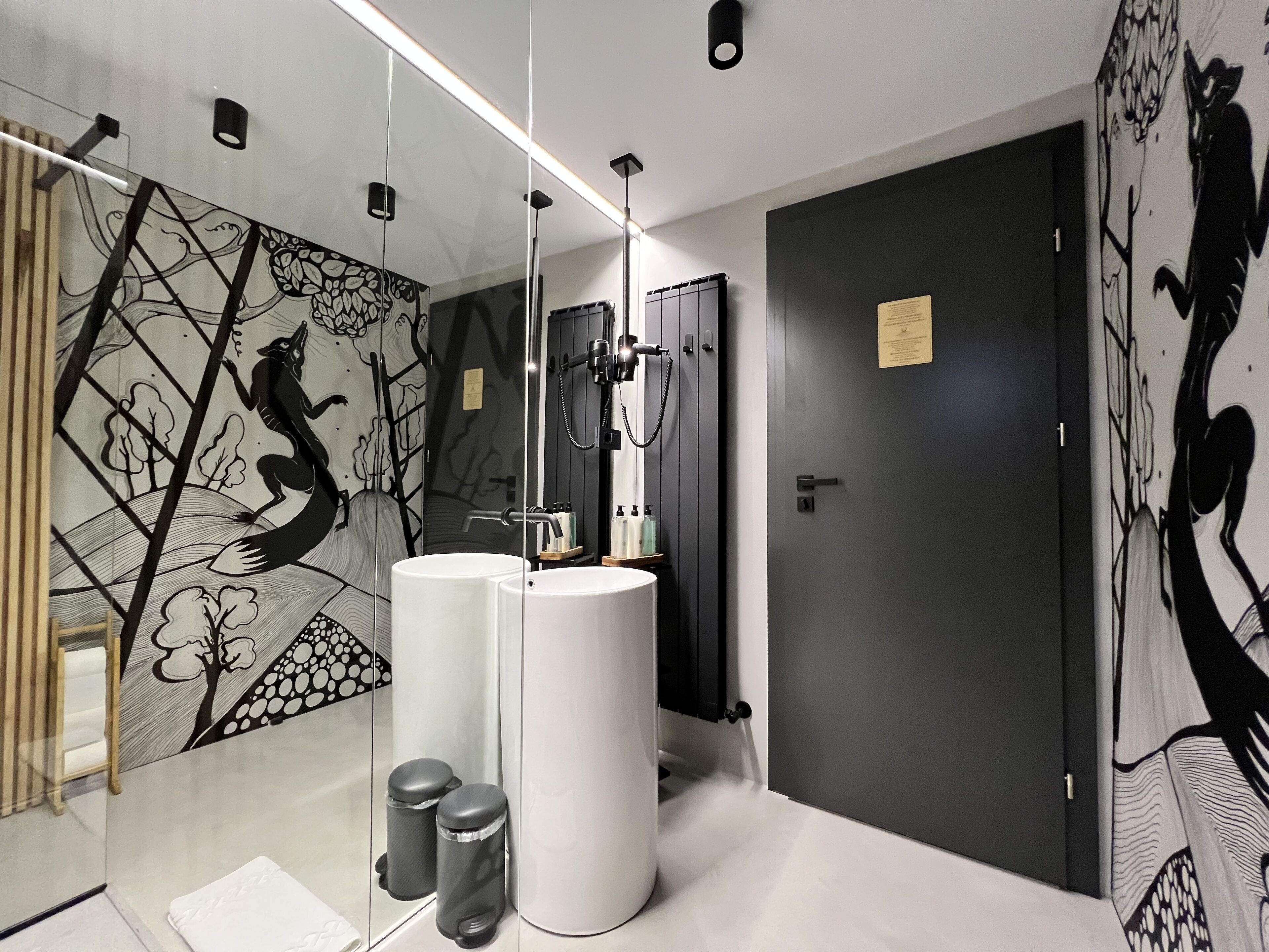 Une salle de bain moderne avec un grand miroir, deux lavabos, une douche et une porte dotée d’un panneau foncé.