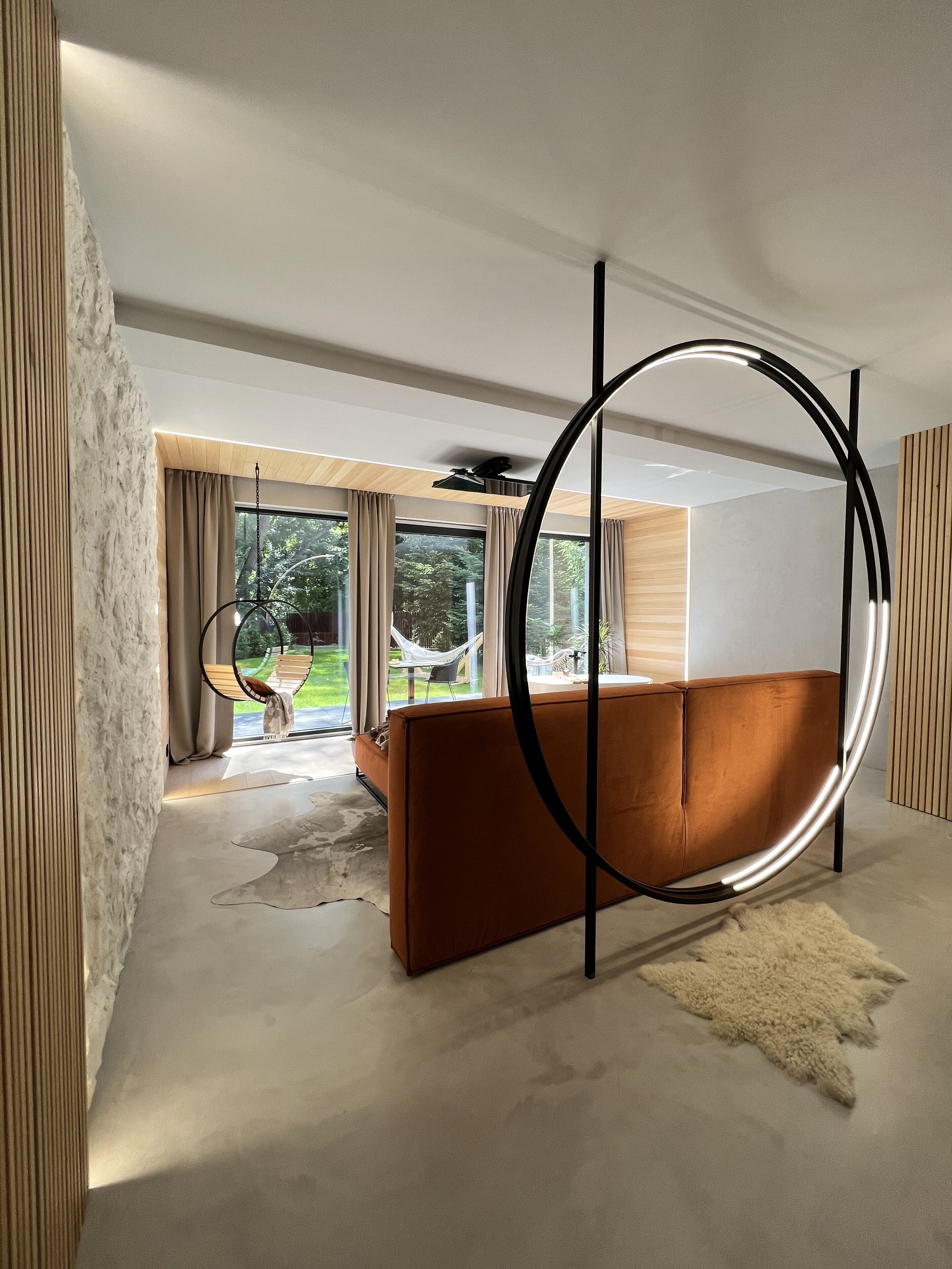 Una sala de estar moderna con una lámpara circular grande, un sofá marrón y vistas a un jardín a través de ventanas amplias.