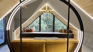 A modern loft bedroom with a sloped ceiling, a circular light fixture, and a view of trees through large windows. - Garden Retreat  (Bran)