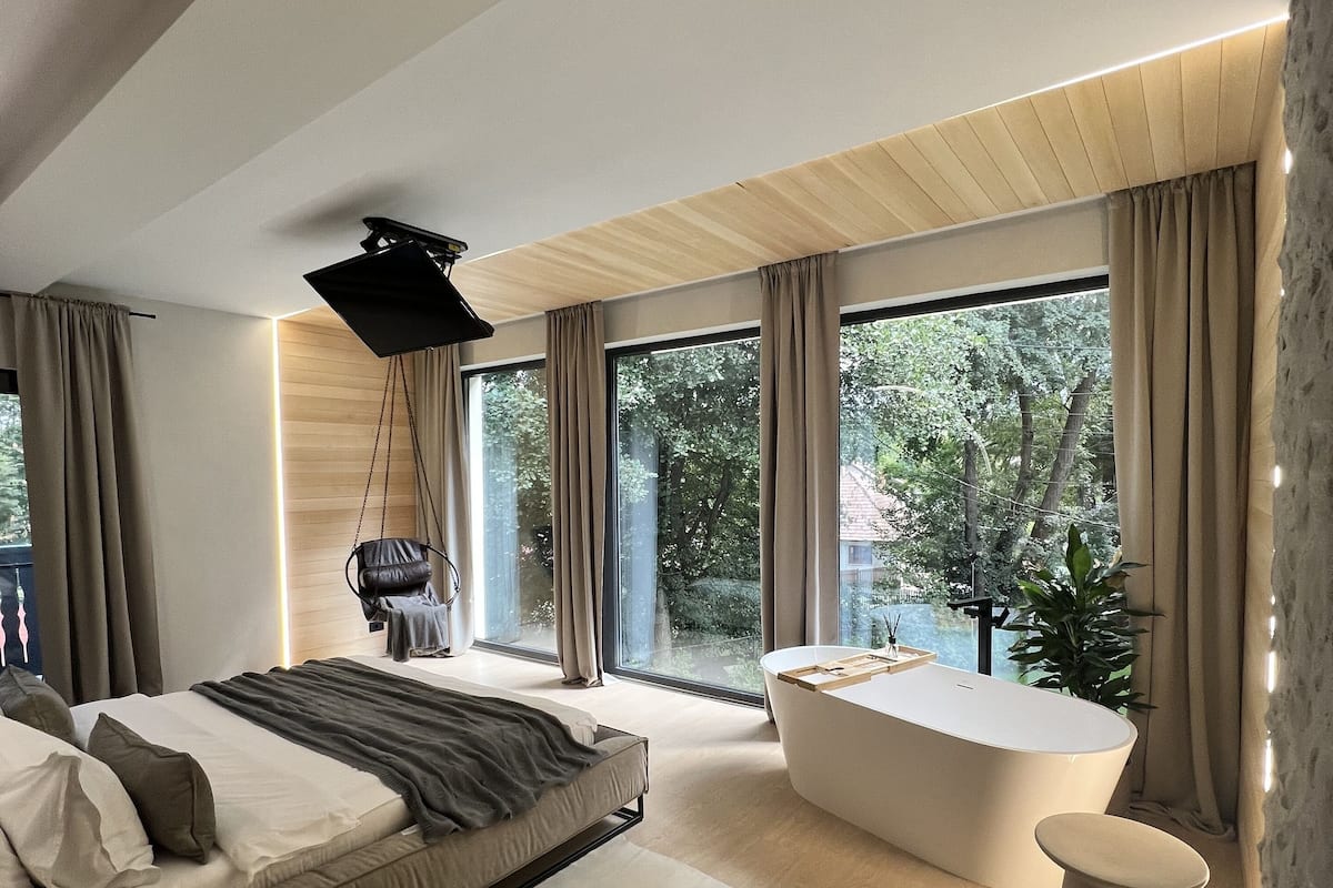 A modern bedroom with a large bed, a bathtub, and a view of trees through large windows.