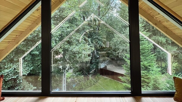 A room with a sloped ceiling and large windows offering a view of a forested area. - Garden Retreat  (Bran)