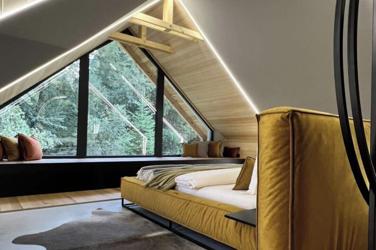 A modern loft bedroom with a sloped ceiling, a large window, a sofa bed, and a unique circular light fixture.