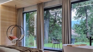 A modern bedroom with a large bed, a hanging chair, a bathtub, and a view of a garden through large windows. - Garden Retreat  (Bran)