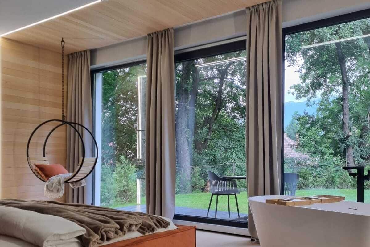 A modern bedroom with a large bed, a hanging chair, a bathtub, and a view of a garden through large windows.