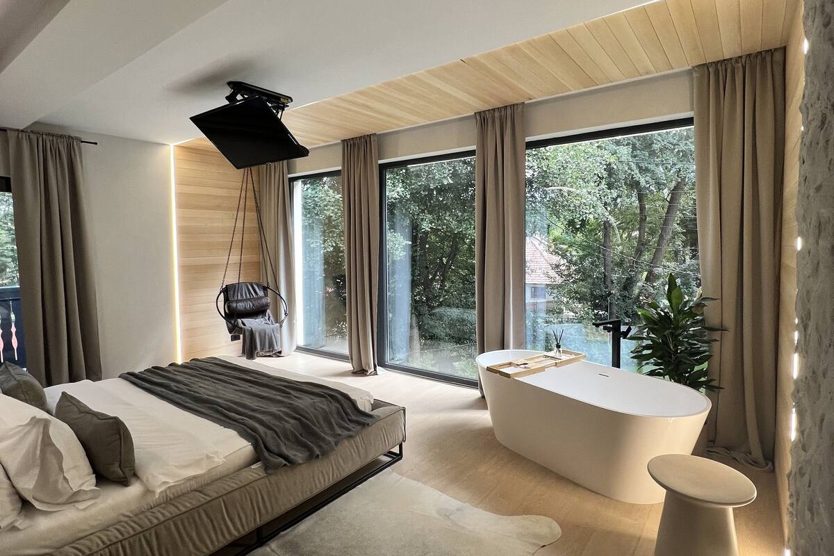 A modern bedroom with a bed, a large window, a bathtub, and a hanging light fixture.