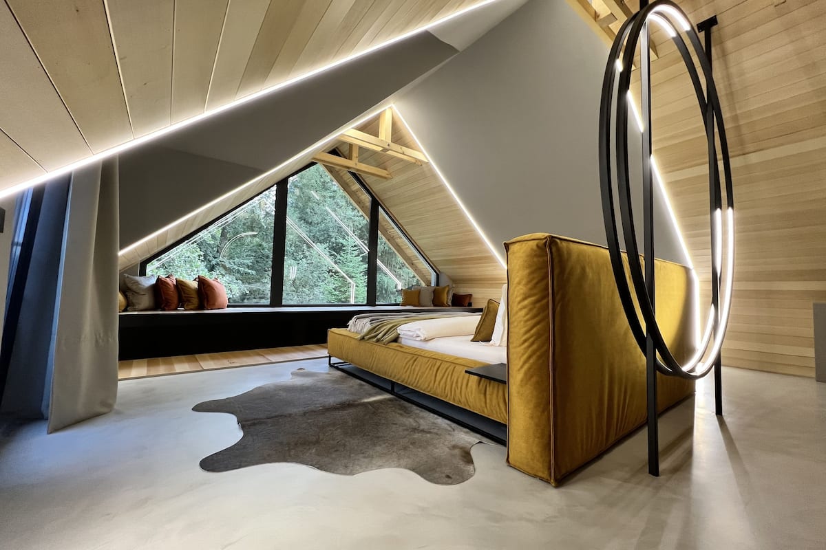 A modern loft bedroom with a sloped ceiling, large windows, a sofa bed, and a circular light fixture.