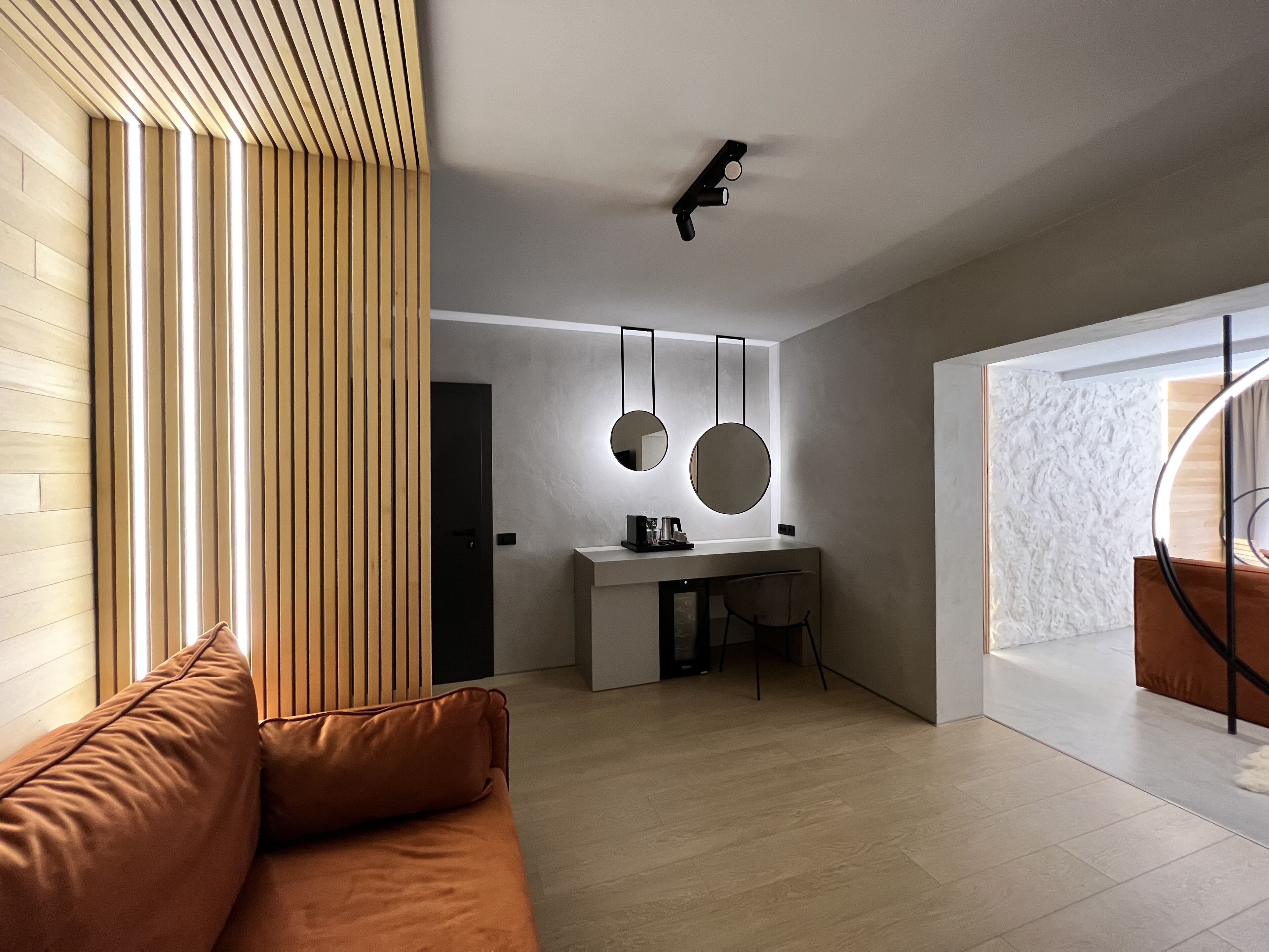 A modern living room with a brown sofa, a wooden slatted wall, a desk with two round mirrors, and a ceiling light fixture.