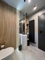 A modern bathroom with a toilet, a sink, and a mirror. There is a wooden slat wall on one side and a dark door on the other.
