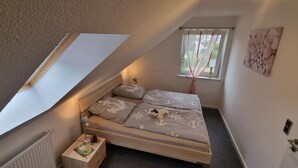 A small bedroom with a sloped ceiling, a single bed, a nightstand, a window with curtains, and a wall-mounted radiator.