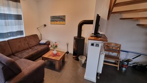 A living room with a brown sofa, a wooden coffee table with a vase of flowers, a small table with a lamp, a painting on the wall, and a wood-burning stove.