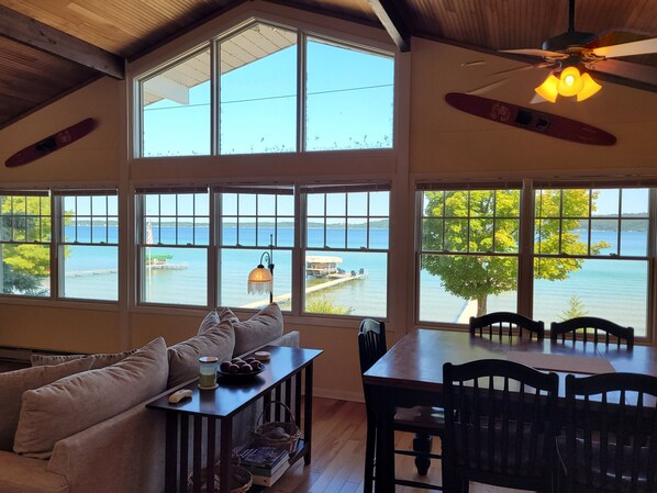 A room with large windows overlooking a body of water, a ceiling fan, and a dining area with chairs and a table.