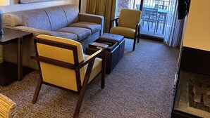 A hotel room with a sofa, armchair, coffee table, and a large painting on the wall. There is a window with curtains and a view of the outside.