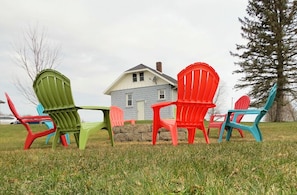 Mobiliario exterior con sillas Adirondack de varios colores, un muro de piedra y una casa al fondo.
