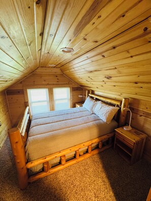 Ein Schlafzimmer einer Holzhütte mit schrägem Dach, einem Doppelbett, einem Nachttisch und einem Fenster.