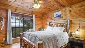 A wooden cabin bedroom with a large bed, a window with curtains, a ceiling fan, and a nightstand with a lamp. - Mountain view cabin near Ellijay w/ hot tub! (Gilmer County)
