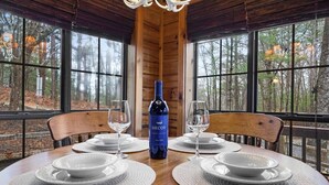 A dining table with four white plates and two wine glasses, a bottle of Decoy wine, and a wooden ceiling with large windows overlooking a forest. - Mountain view cabin near Ellijay w/ hot tub! (Gilmer County)