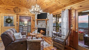 A cozy cabin interior with a stone fireplace, wooden furniture, and a large window overlooking a scenic view. - Mountain view cabin near Ellijay w/ hot tub! (Gilmer County)
