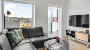 A modern living room with a leather sofa, a flat-screen TV, and a large window offering a view of a residential area. - Amazing apartment in Ringkøbing (Ringkøbing)