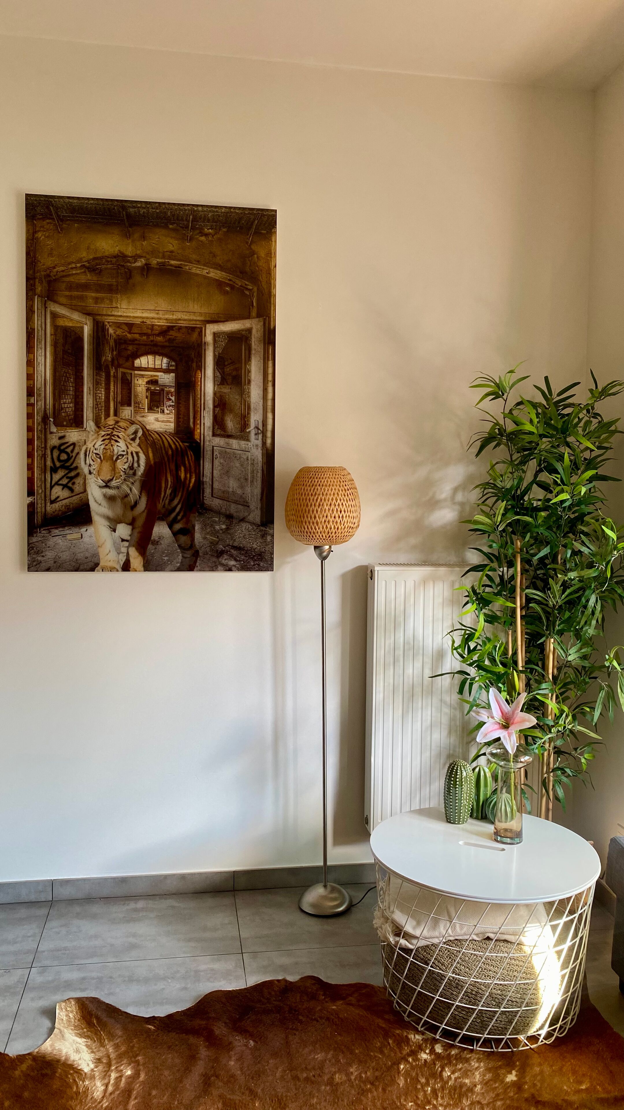 Une pièce décorée d’un tableau représentant un tigre, avec un lampadaire, une plante en pot, un vase rempli de fleurs et une table ronde sur laquelle se trouve un panier en fil de fer.