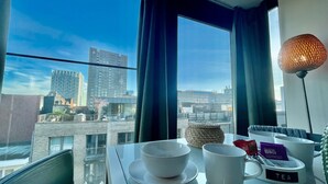 Habitación de hotel moderna con vista a la ciudad, una mesa de comedor con tazas de té y café, y una lámpara.