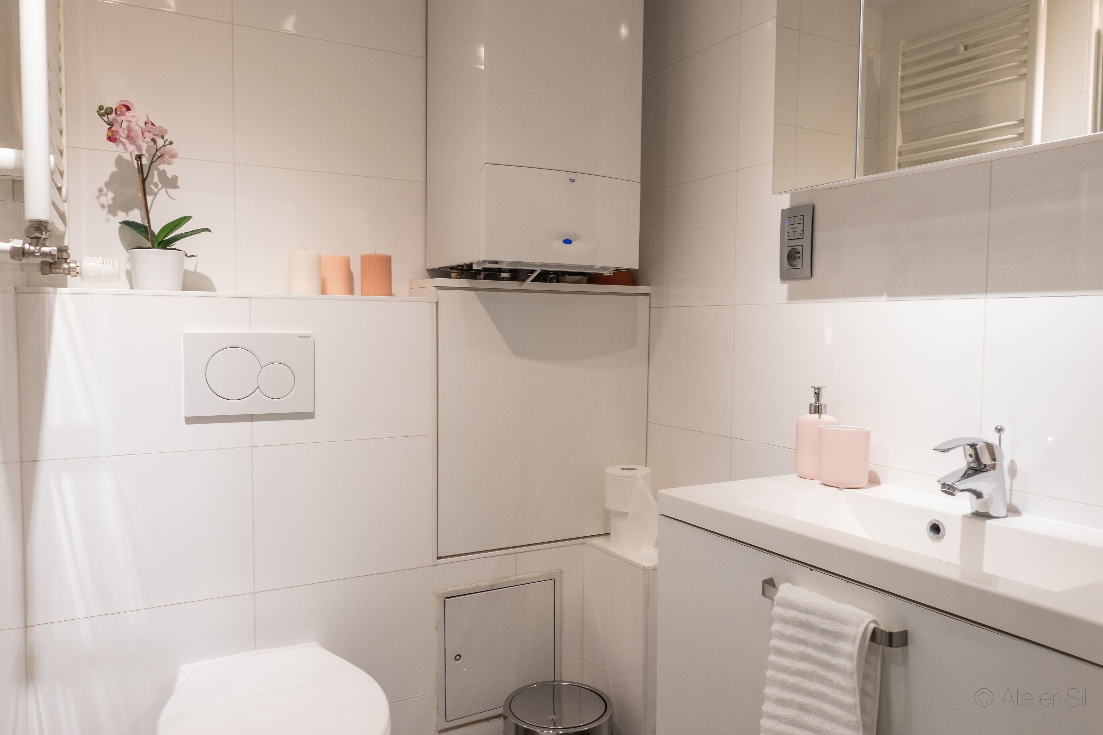 A modern bathroom with a toilet, sink, and shower. There is a potted orchid on the sink counter, a towel rack, and a trash can.