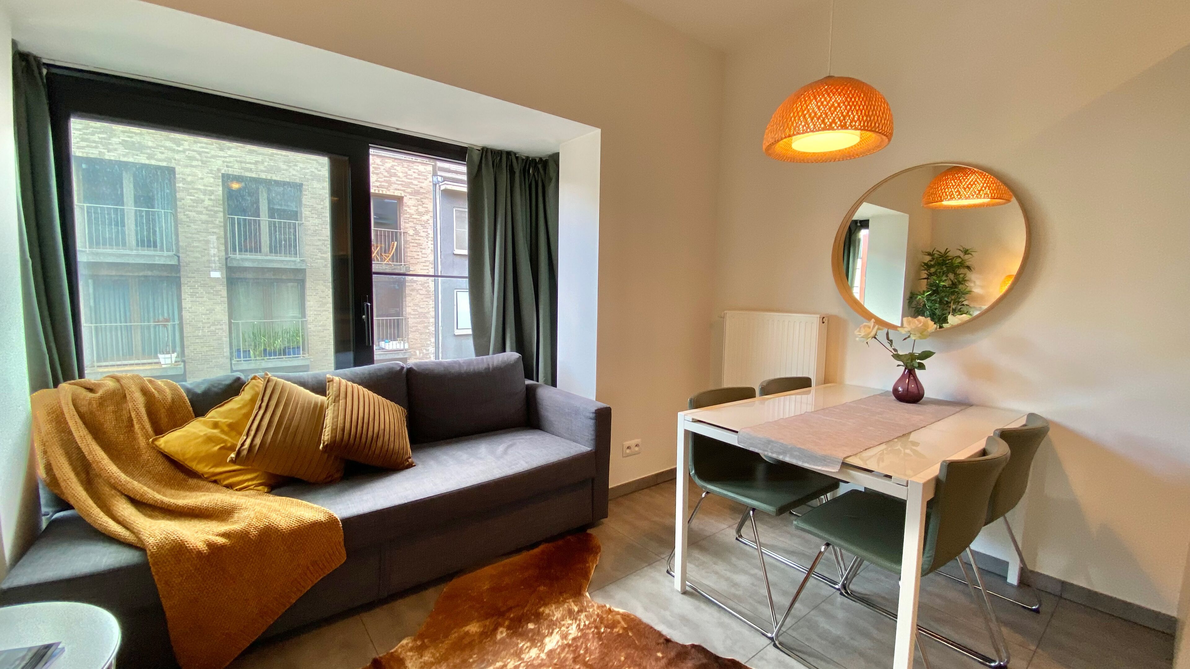Una habitación moderna con un sofá gris, una mesa de comedor con sillas, un espejo y un ventanal con cortinas.