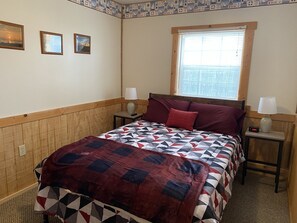 Une chambre avec une tête de lit en bois, une couvre-lit matelassé, deux lampes de chevet, une horloge numérique et une fenêtre avec des stores.