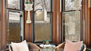 A corner room with two wicker chairs, a small round table, and a view of trees through large windows.